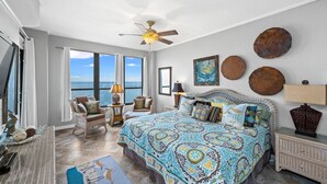 Een ruime slaapkamer met een groot bed, twee stoelen, een plafondventilator en uitzicht op de oceaan.