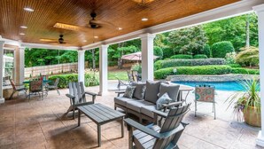 Un patio techado con mobiliario exterior, piscina y vistas a un jardín paisajístico.