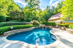 Une piscine bordée de pierres, entourée d’une végétation luxuriante, avec une maison en arrière-plan.