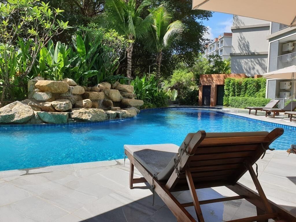 Una piscina con una tumbona de madera, rodeada de plantas tropicales y un muro de piedra.