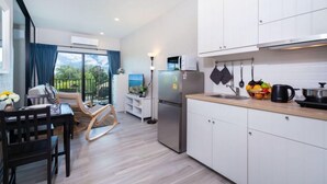 A modern kitchen with white cabinets, a stainless steel refrigerator, and a microwave. There is a dining area with a table and chairs, and a view of greenery outside. - The Title Naiyang Residence (Sa Khu)