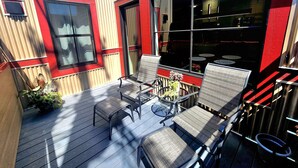 Un espacio de estar al aire libre cubierto, con sillas de metal y una pequeña mesa, rodeado por un edificio de color rojo y beige con ventanas amplias.