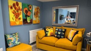 A room with a yellow sofa, a matching ottoman, and a colorful abstract pillow. There are two paintings on the wall, one with sunflowers and another with a floral pattern. - Fabulous apartment for 2 in Llansteffan (Llansteffan)