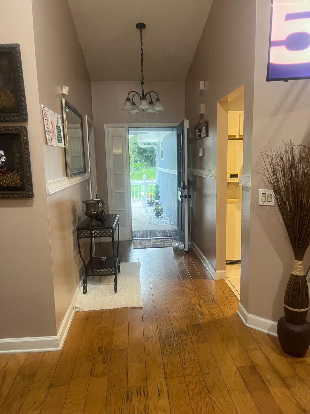 A hallway with a wooden floor, a chandelier, a door leading outside, and a wall-mounted light fixture.