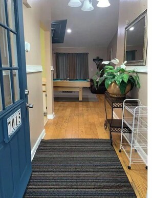 Enter a hallway leading to a room with a pool table, featuring a door with a number plate, a striped rug, and a plant on a metal rack.