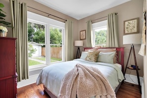Un dormitorio con una cama grande, dos ventanas con cortinas, una lámpara de noche y suelo de madera.