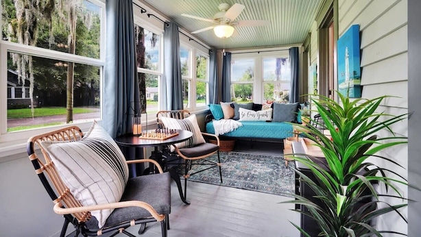 A cozy room with a ceiling fan, a sofa with pillows, a table with chess, and a large window with curtains. - Tranquil Avondale Home, Walkable to Food & Shops! (Jacksonville)