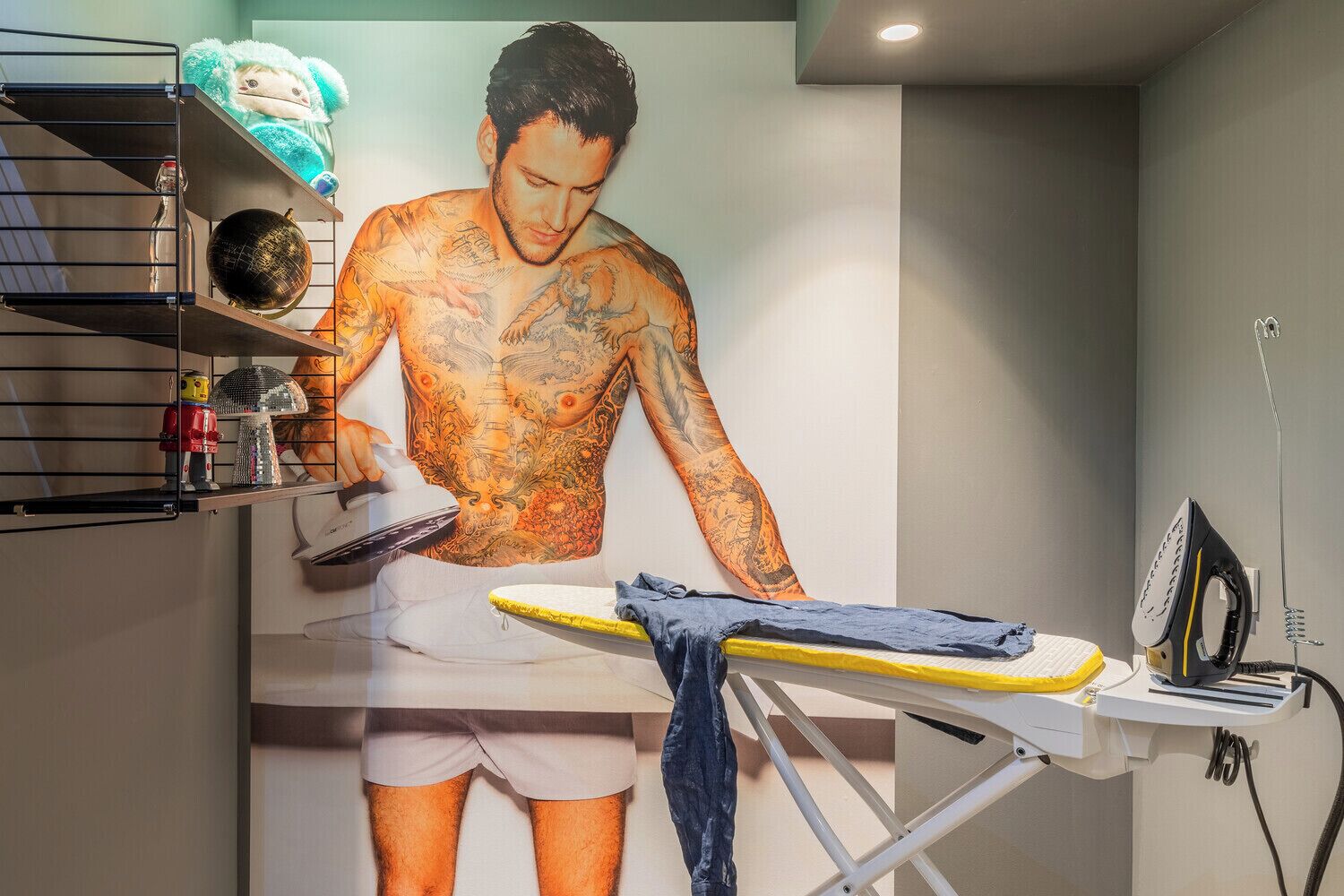 Una habitación con un gran mural de un hombre sin camisa planchando ropa, una estantería con varios objetos y una tabla de planchar.