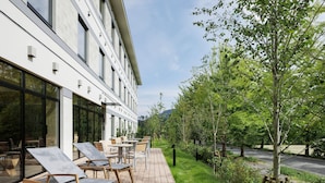 An outdoor seating area with wooden tables and chairs, a paved walkway, and a building with large windows and a modern design.