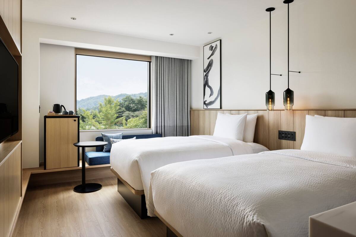 A hotel room with two beds, a small table, a TV, and a window with a view of greenery.