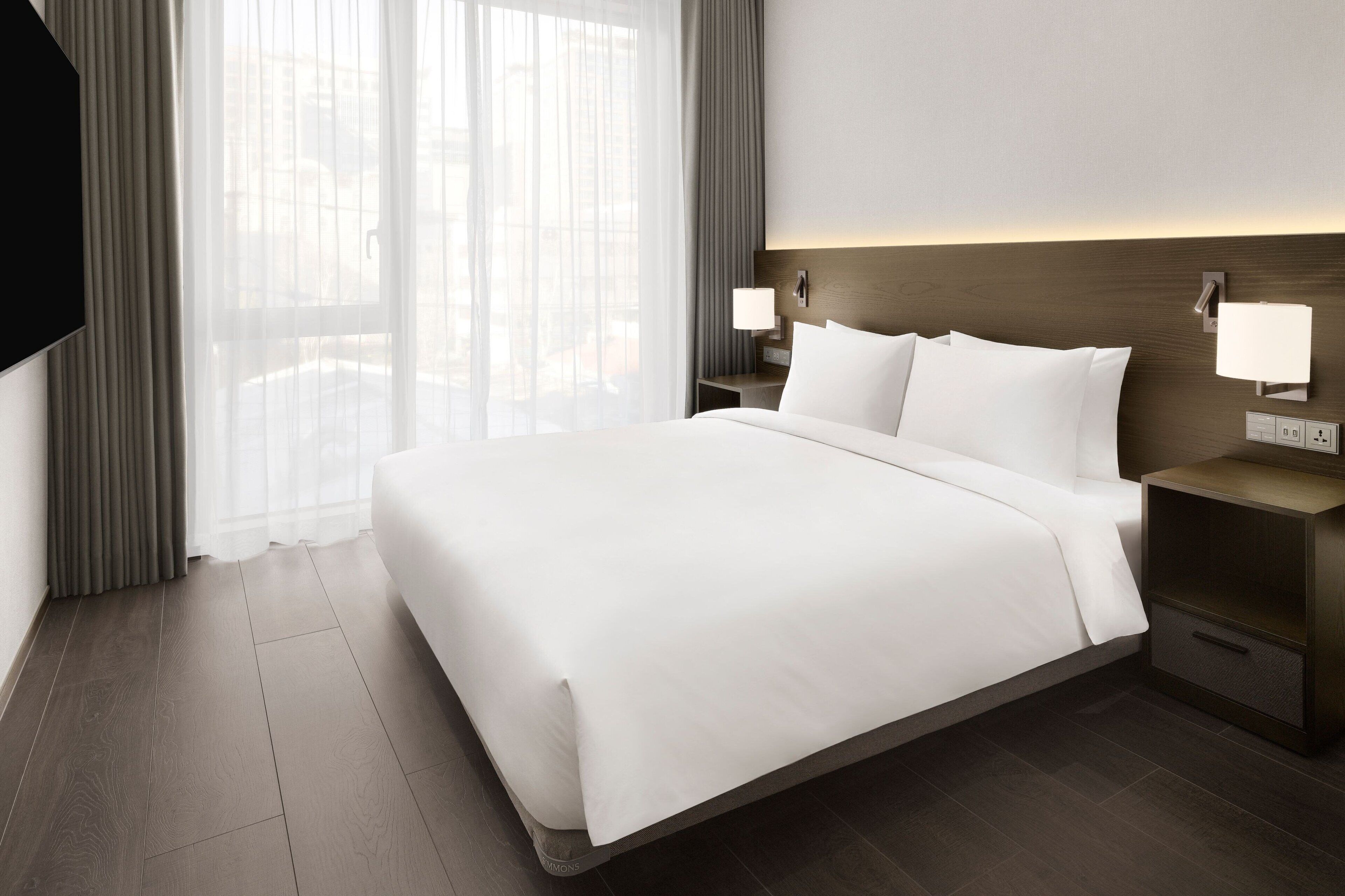 Een moderne hotelkamer met een groot bed, twee nachtkastjes met lampen en uitzicht door dunne gordijnen.