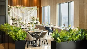 A modern restaurant with a city view, featuring a central stage with floral decorations, multiple dining tables, and lush green plants in the foreground.