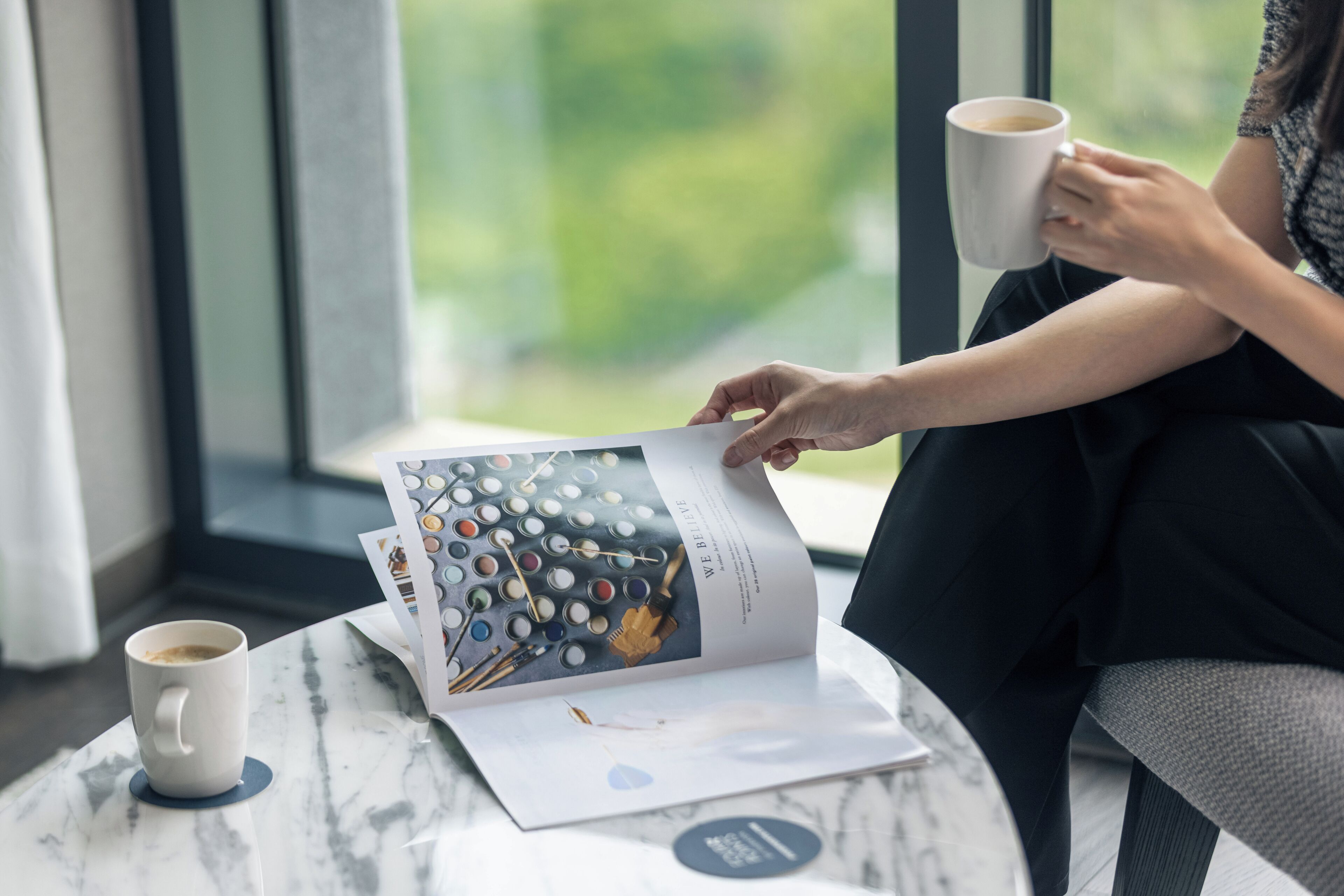 A person reading a magazine with a coffee cup on a table by a window.