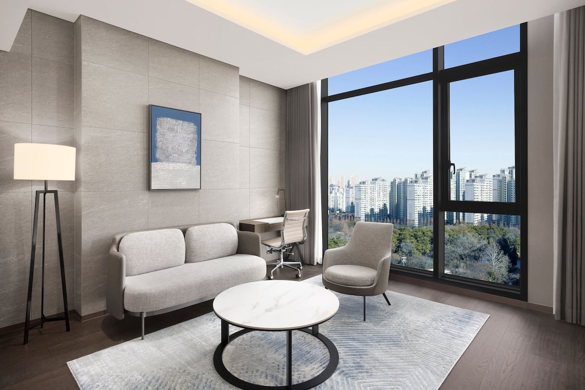 A modern living room with a sofa, armchair, and coffee table, a floor lamp, and a large window with a city view.