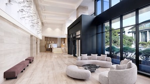 A spacious lobby with a modern design, featuring a curved seating area, a staircase in the background, and large windows offering a view of the outdoors.