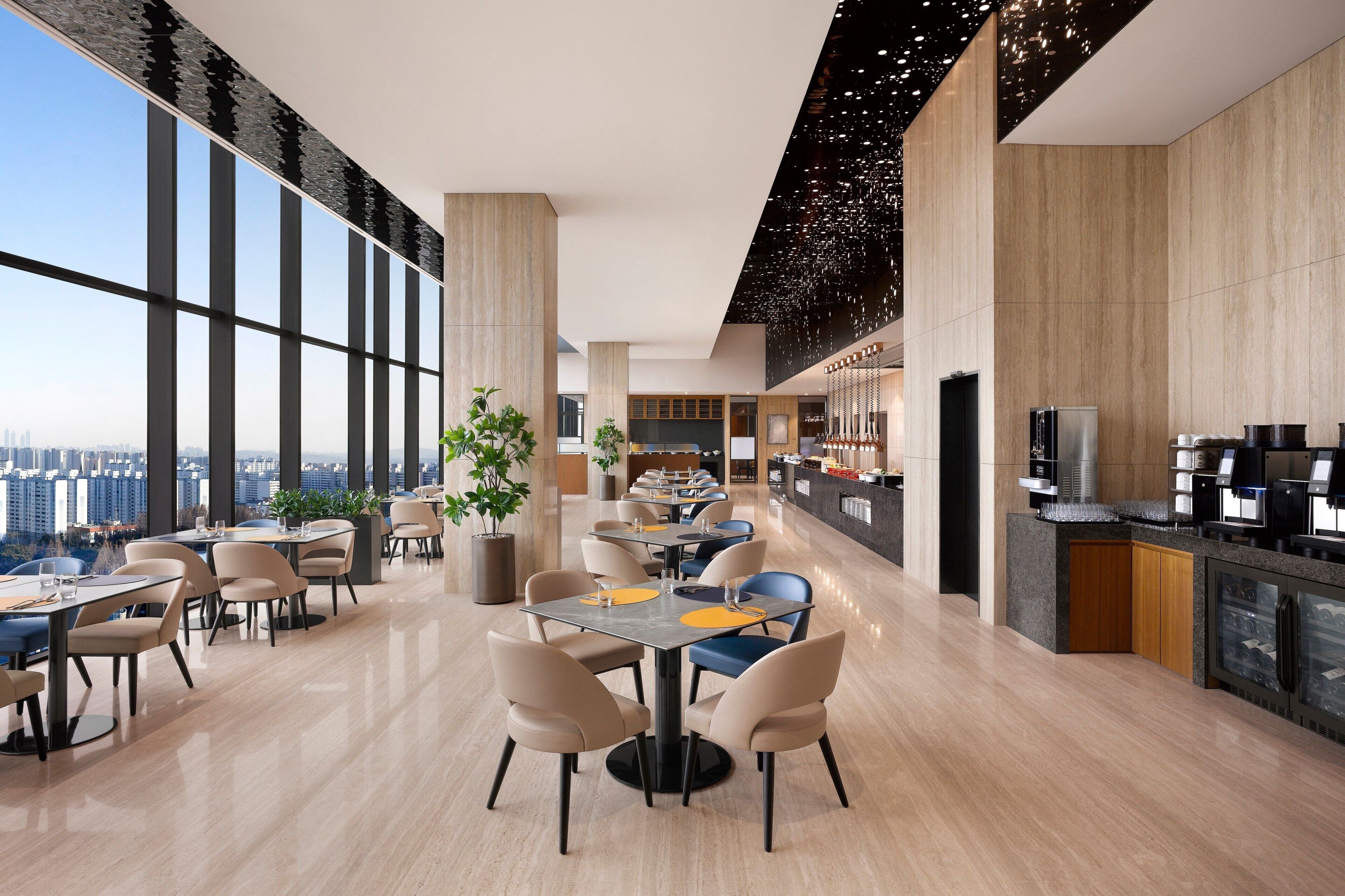 A modern dining area with high ceilings, large windows, and a city view.