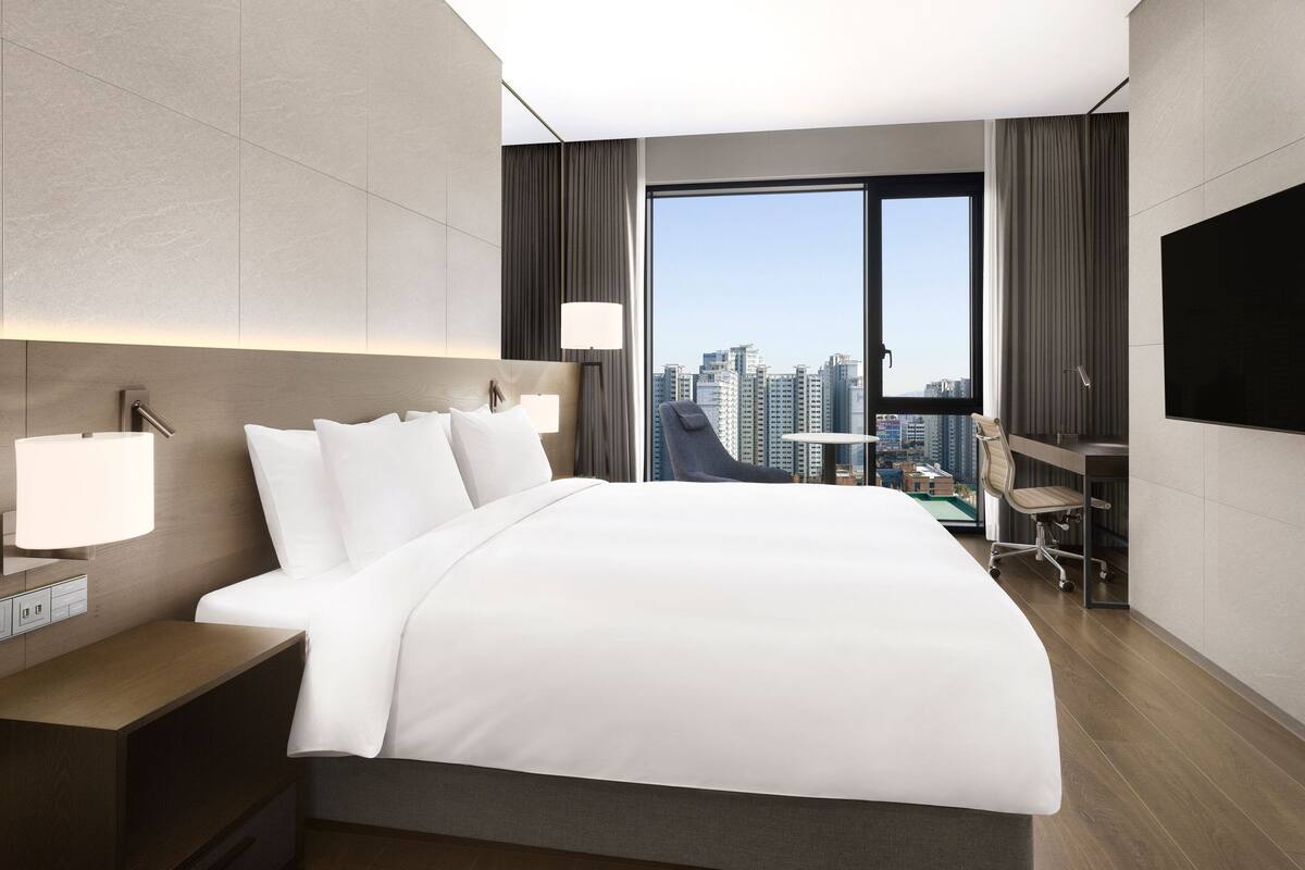A modern hotel room with a large bed, a desk, and a chair, a TV mounted on the wall, and a city view through a large window.