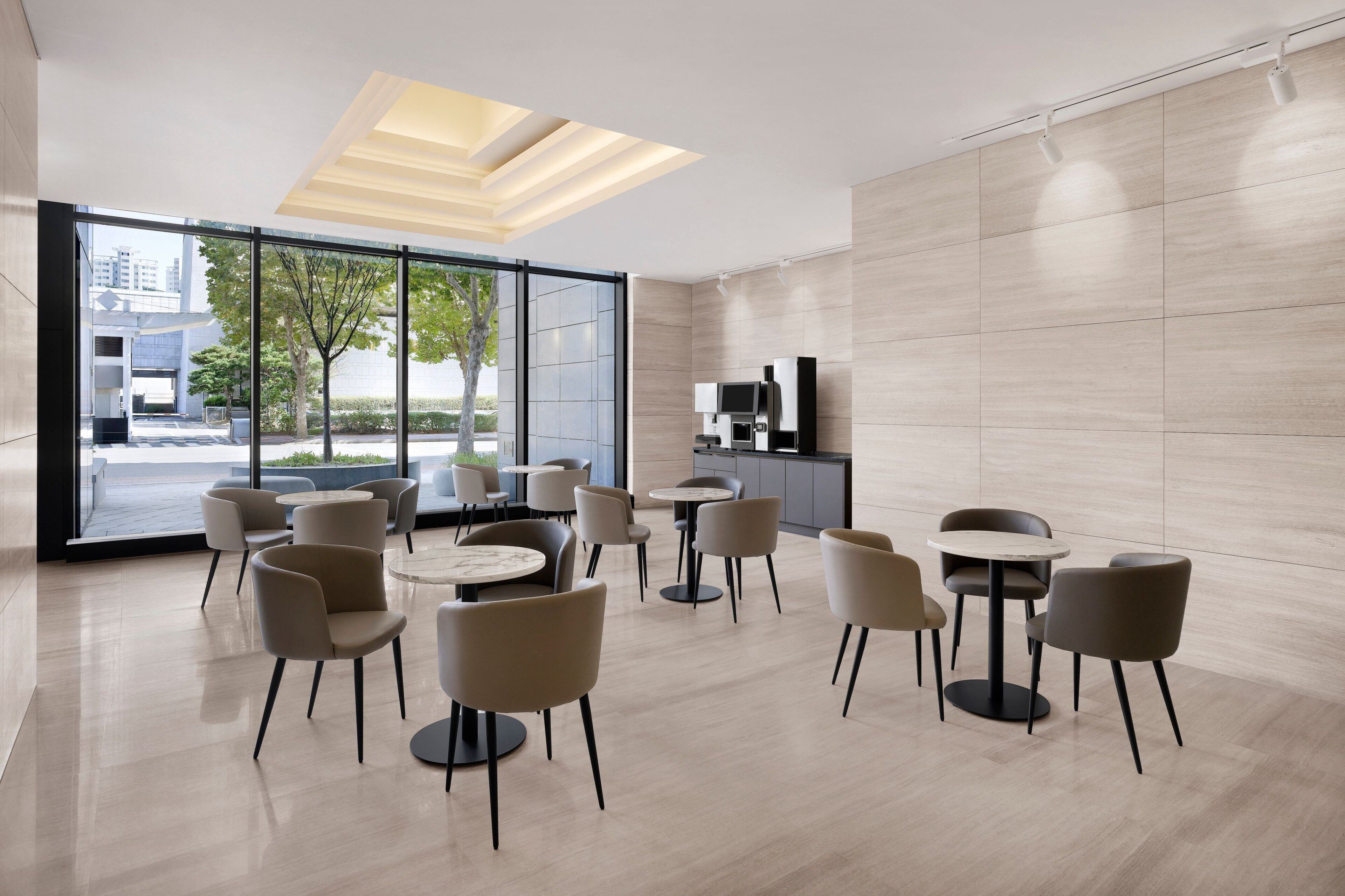 A modern cafe with round tables and chairs, large windows, and a kitchen area.