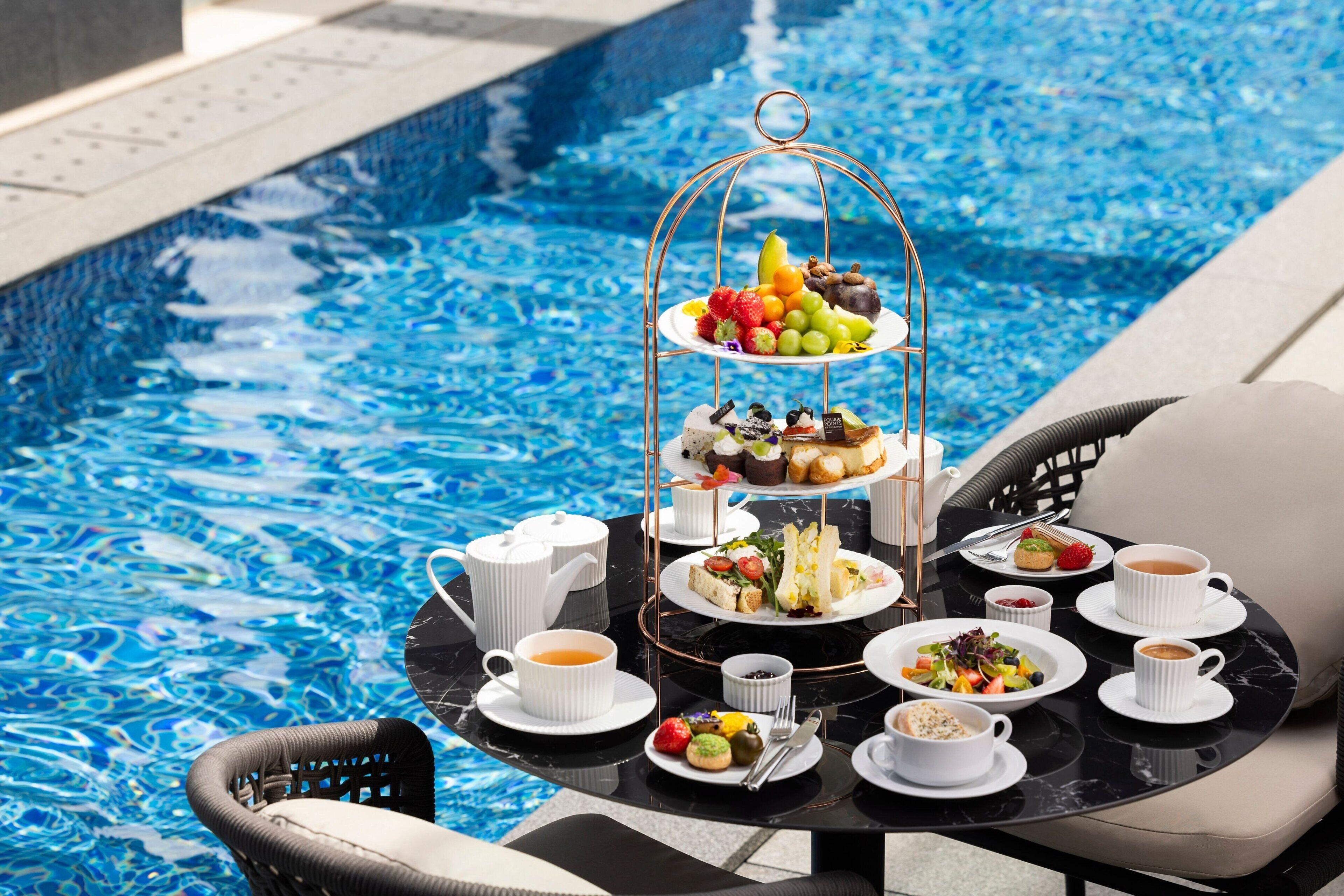 Mesa junto a la piscina con un soporte de postres de tres niveles, diferentes platos de comida, vasos de bebidas y una silla de mimbre.