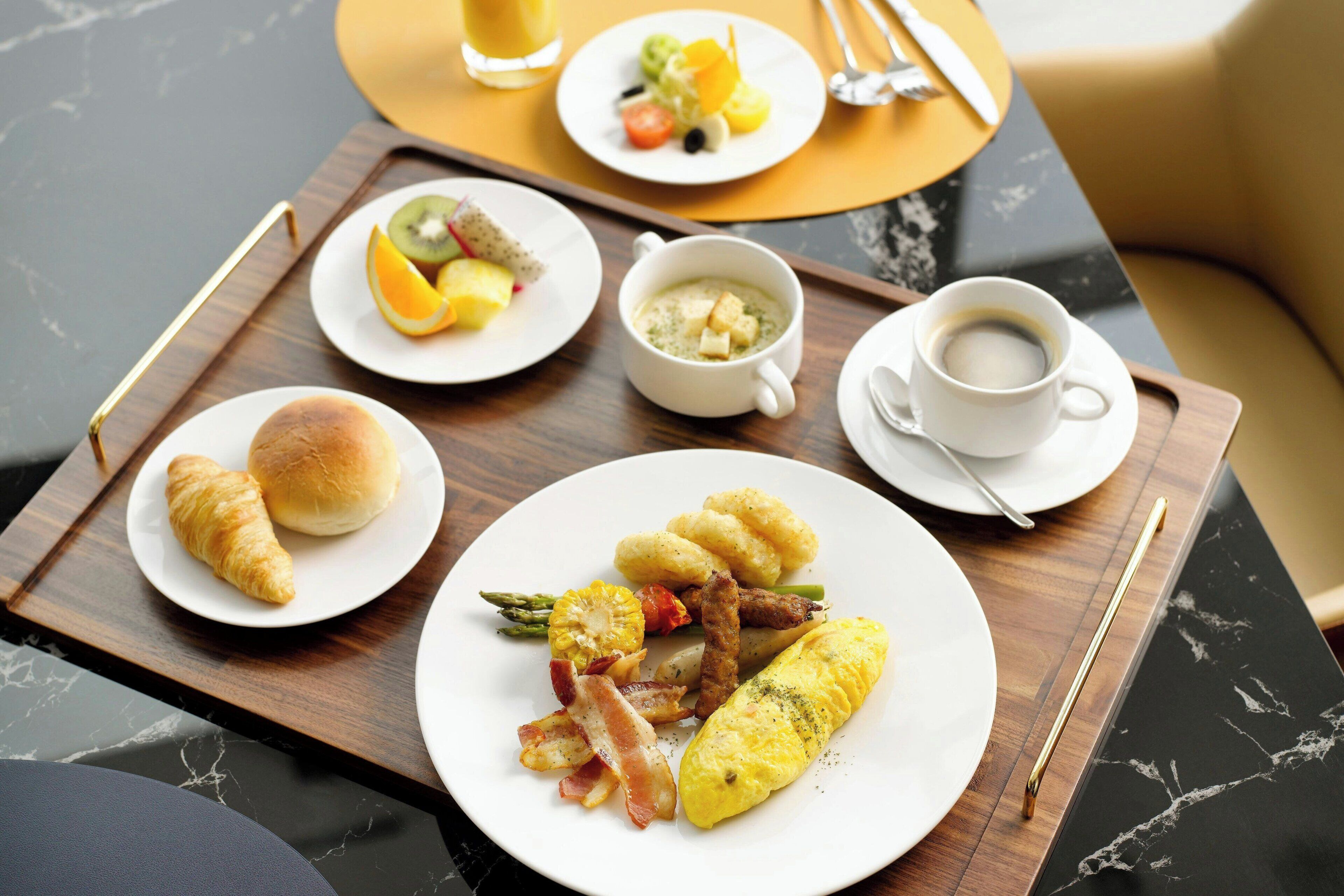 Ein Frühstücksbrett mit Croissants, einem Omelett, Spargel, Speck und einer Tasse Kaffee auf einer Marmortischplatte.
