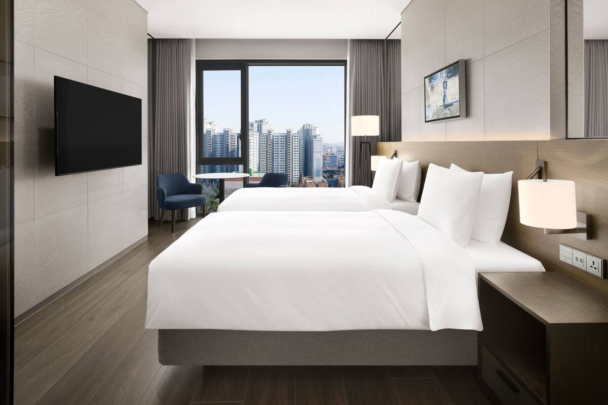 A hotel room with a large bed, a TV, a desk with a lamp, and a view of the cityscape.