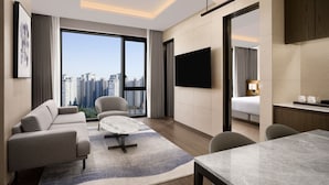 A modern hotel room with a large window offering a city view, a flat-screen TV mounted on the wall, a sofa, a dining table, and a small kitchen area.