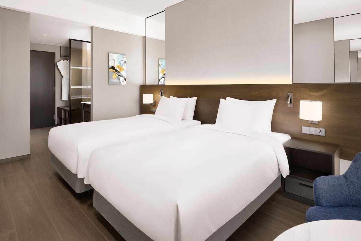 A hotel room with two beds, a nightstand with a lamp, a chair, and a mirror on the wall.