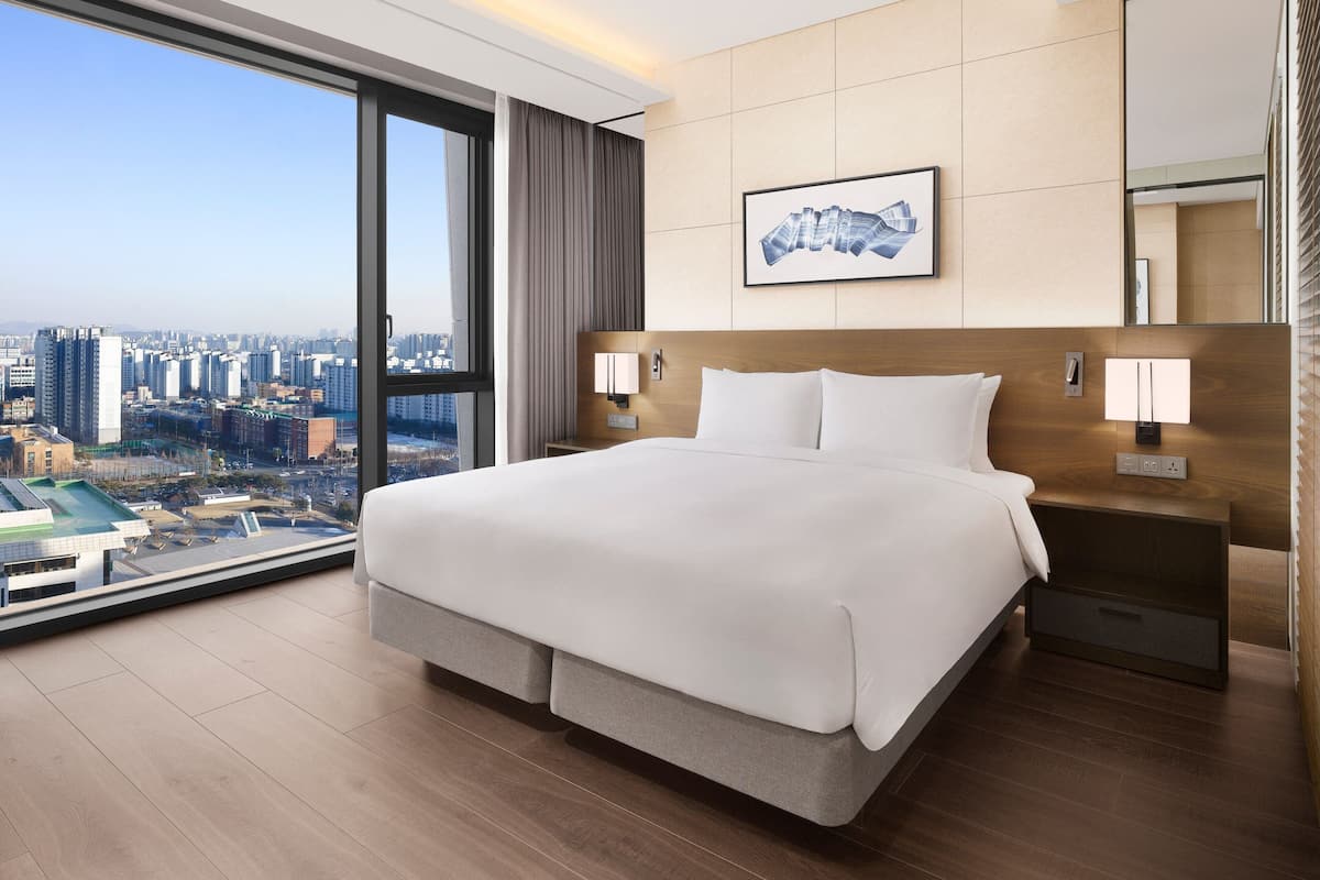 A modern hotel room with a large bed, a city view, and a framed artwork on the wall.