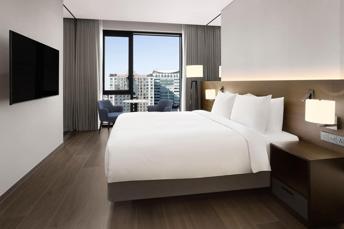 A modern hotel room with a large bed, a flat-screen TV, a desk with a lamp, and a view of the city through large windows.