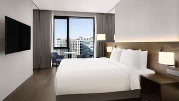 A modern hotel room with a large bed, a flat-screen TV, a desk with a lamp, and a view of the city through large windows.
