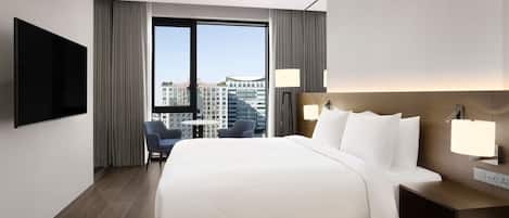Una habitación de hotel moderna con una cama grande, un televisor de pantalla plana, un escritorio con lámpara y vistas a la ciudad a través de amplios ventanales.