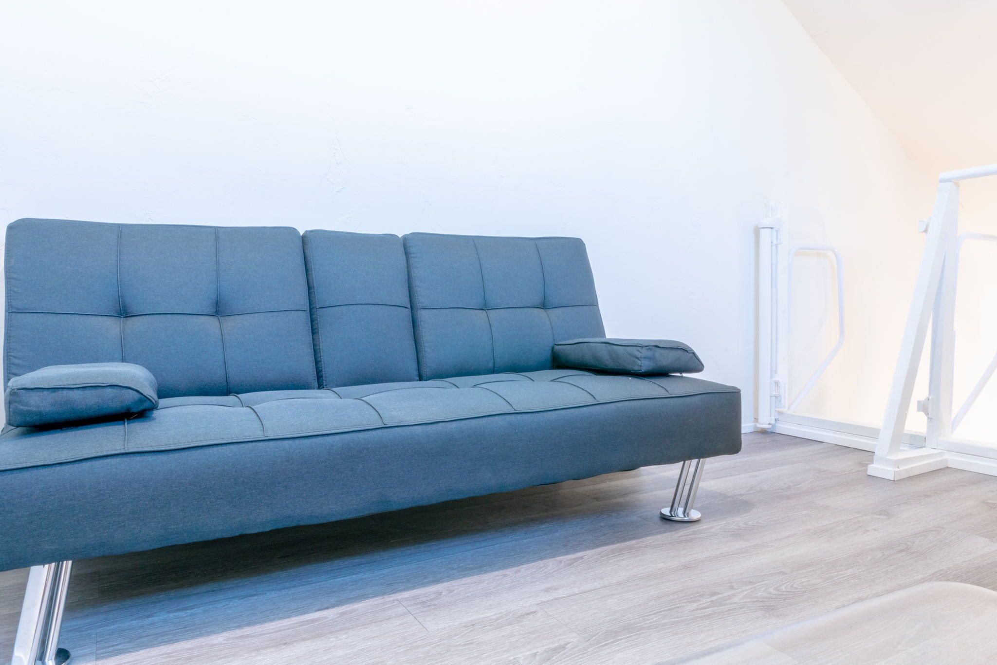 A modern grey sofa with a cushion, positioned against a white wall in a room with wooden flooring.