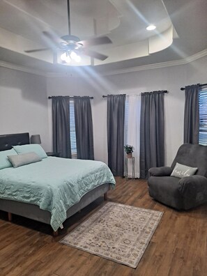 Ein Schlafzimmer mit einem Bett, einem grauen Sessel, einem kleinen Tisch mit Pflanze und einem Deckenventilator.