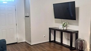 A living room with a dark-colored sofa, a small console table with a clock, and a flat-screen TV mounted on the wall. - Beautiful house close to the sea (Corpus Christi)