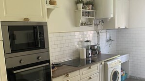 Een moderne keuken met witte kastjes, een ingebouwde oven, magnetron en vaatwasser. Er is een houten aanrechtblad, een hanglamp en een klein plankje met planten.