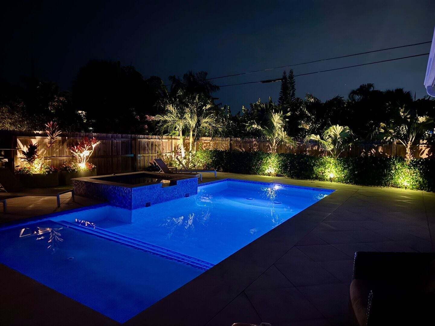 A swimming pool with a tiled deck, surrounded by a wooden fence and illuminated plants.
