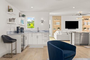 A modern kitchen with white cabinets, a marble countertop, and a built-in oven. There is a dining area with a white sofa and a blue chair.