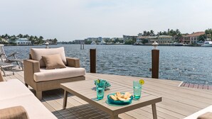 A deck with a sofa, a table with drinks and snacks, and a view of the water and buildings in the background.