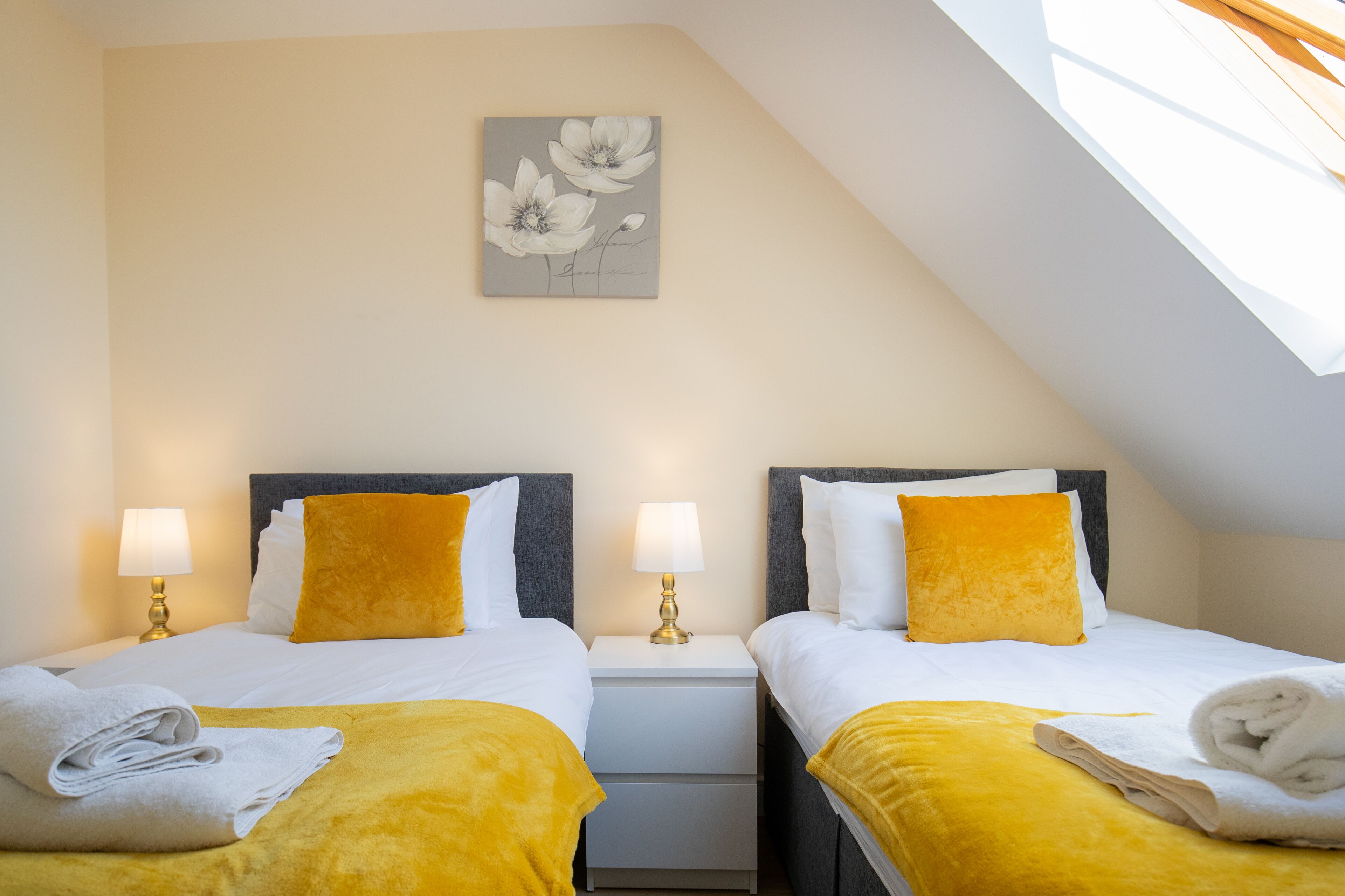 Een hotelkamer met twee bedden, elk met een wit en geel sprei, een nachtkastje met een lamp, en een bloemen schilderij aan de muur.
