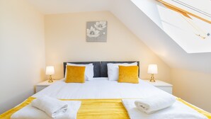 Un lit bien fait, avec une literie blanche et jaune, deux lampes de chevet et une œuvre d’art accrochée au mur.