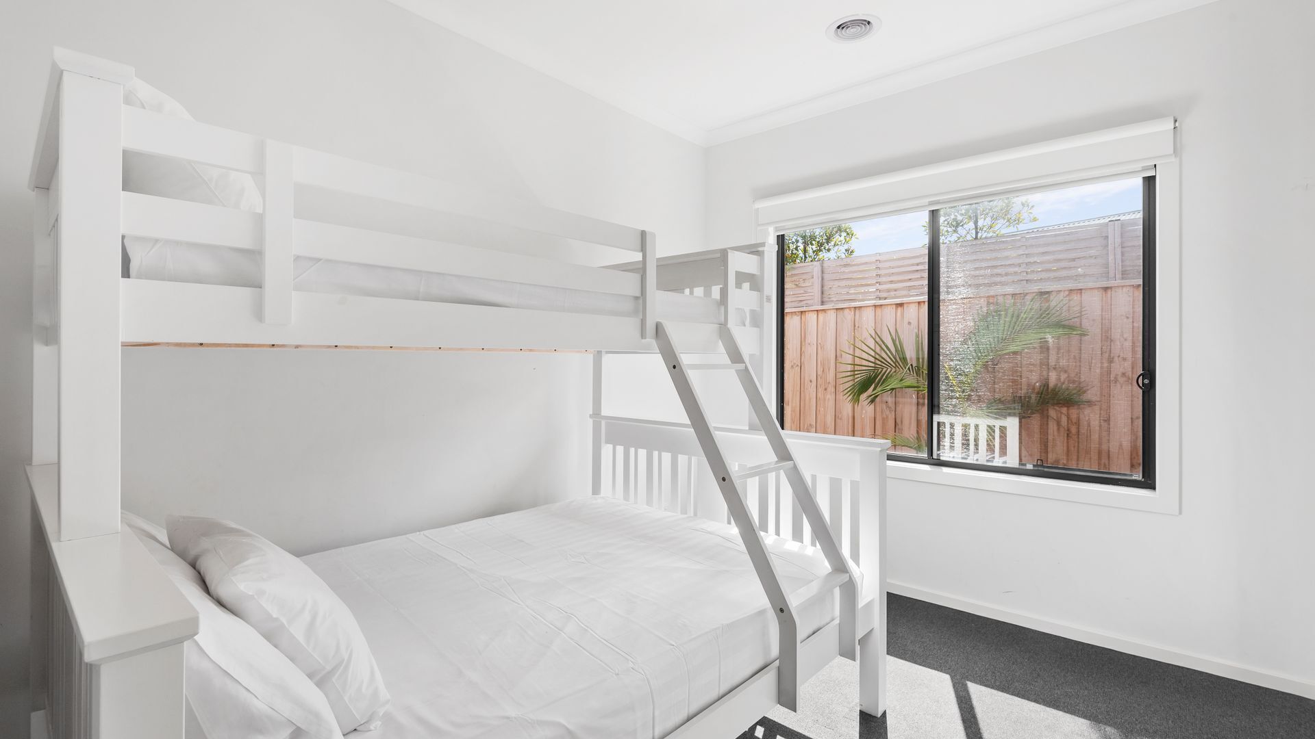 A bunk bed with a ladder, white bedding, and a window with a view of a wooden fence and plants.