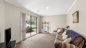A spacious living room with a large sliding glass door leading to a balcony, a flat-screen TV, a striped sofa, and a wooden bookshelf.