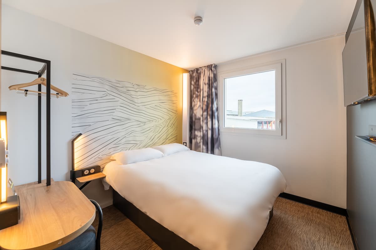 Quarto de hotel com uma cama grande, mesa de cabeceira, luminária na parede, cabideiro e janela com vista para edifícios.
