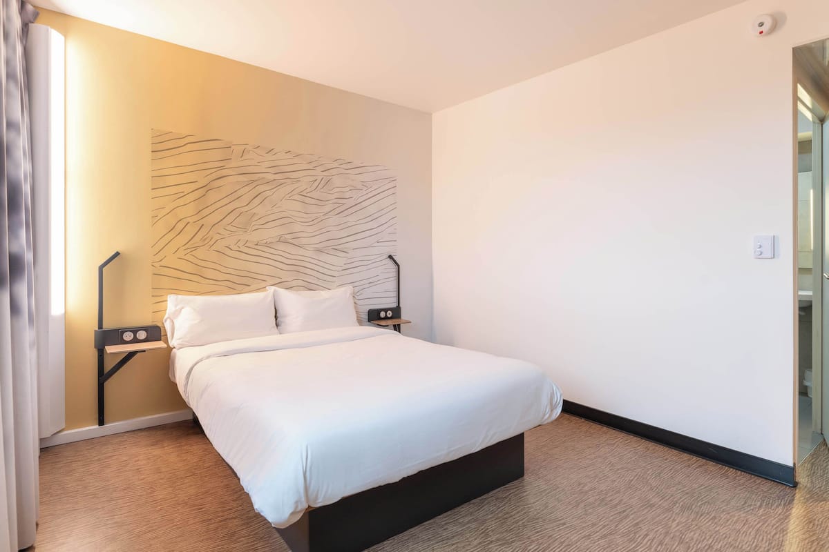 Quarto de hotel moderno com uma cama grande, uma mesa de cabeceira, uma luminária na parede e piso de madeira.