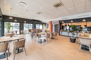 Ein modernes Restaurant mit Holztischen und -stühlen, großen Fenstern und einer Theke mit einer Pflanze.