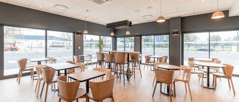 A spacious indoor area with multiple tables and chairs, large windows offering views of the outside, and a modern ceiling design.
