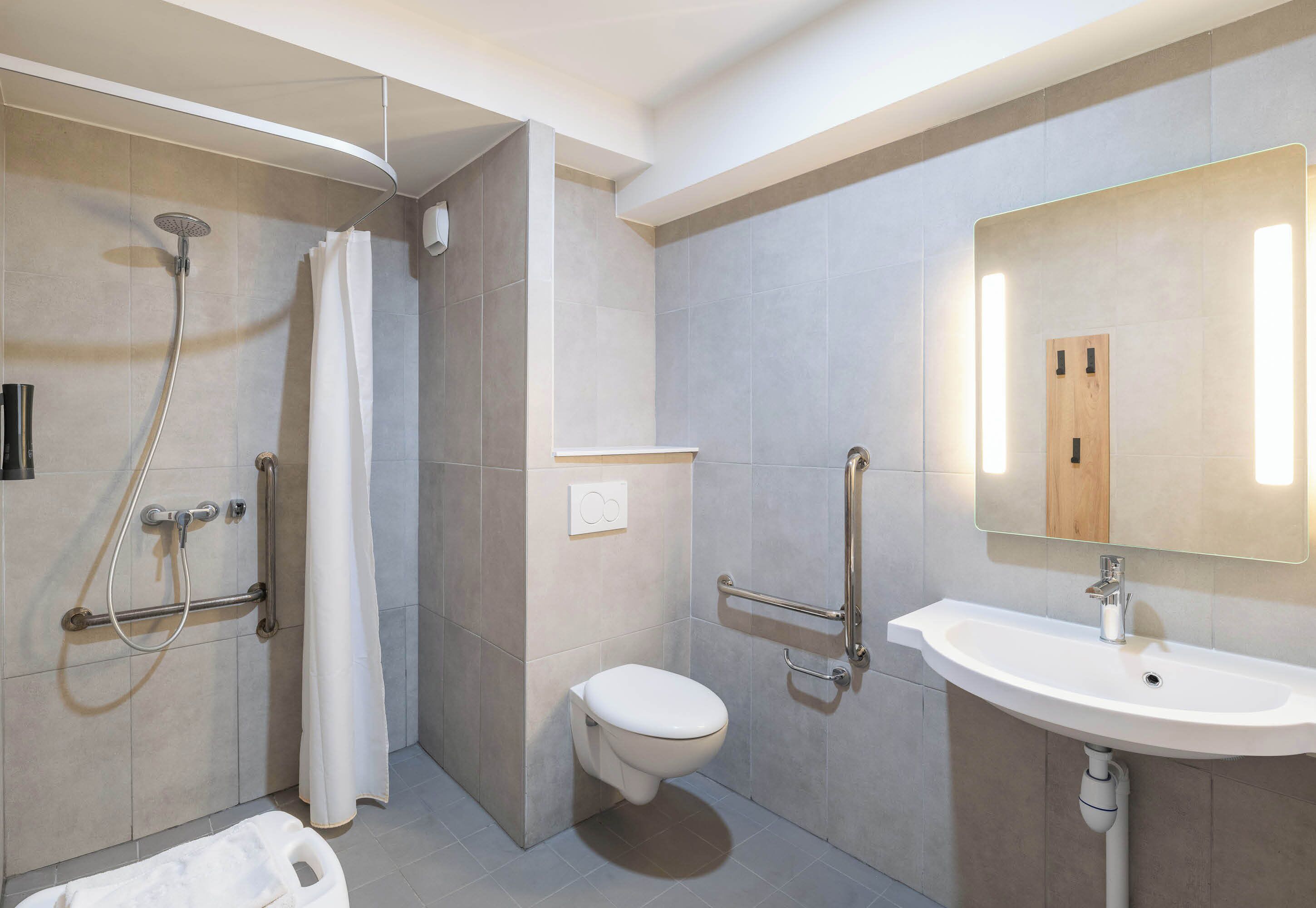 A modern bathroom with a shower, toilet, and sink, equipped with grab bars and a mirror.