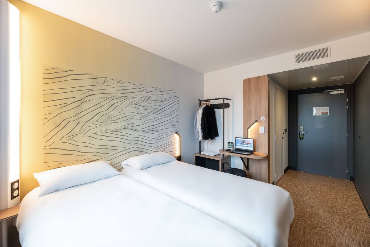Quarto de hotel com uma cama grande, uma escrivaninha com computador, uma obra de arte na parede e uma porta à direita.