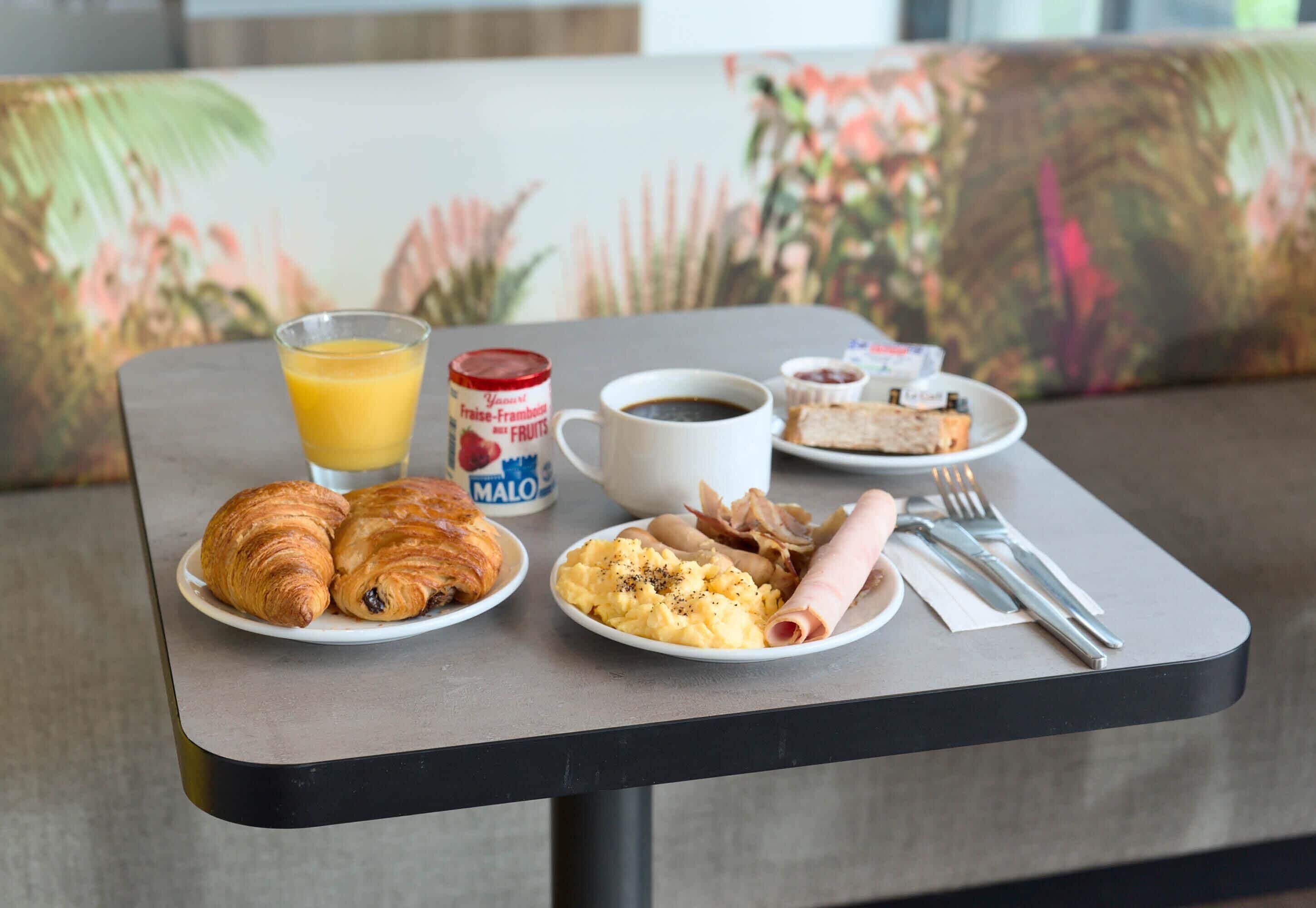 A breakfast table with croissants, scrambled eggs, ham, a glass of orange juice, a cup of coffee, and a can of fruit yogurt.