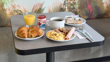 A breakfast table with croissants, scrambled eggs, ham, a glass of orange juice, a cup of coffee, and a can of fruit yogurt.